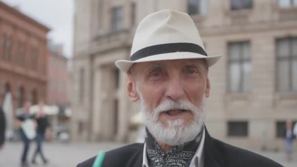 Muž s šedými vousy a modrýma očima mluví proti budovám — Stock video