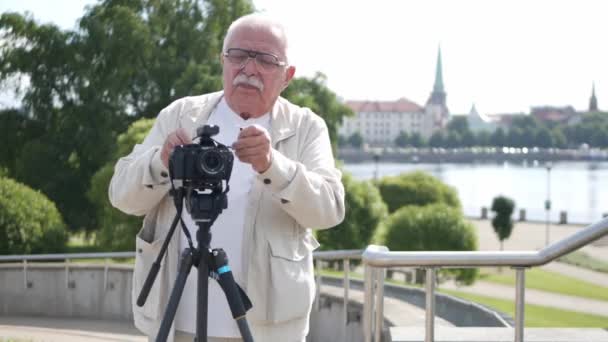 Homem sênior ajusta câmera contra edifícios do rio e da cidade Videoclipe