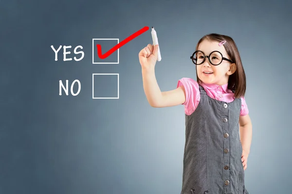 Schattig klein meisje dragen Business jurk en het tekenen van ja in een whiteboard. Blauwe achtergrond. — Stockfoto