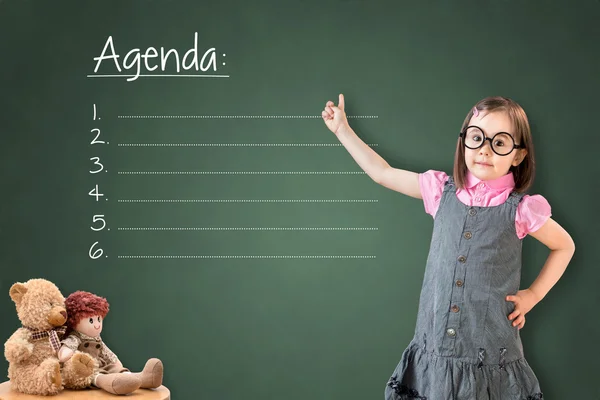 Carino bambina che indossa abito da lavoro e mostrando lista agenda vuota sul bordo gesso verde . — Foto Stock