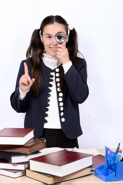 Schoolgirl behind a desk with books looking into a magnifying glass, increasing eye — Stock Photo, Image