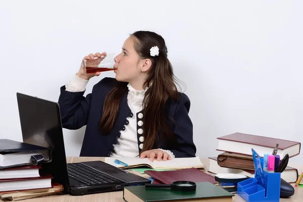Teen schoolgirl at a school desk with a computer and books is drinking a glass of beverage — Stock Photo, Image