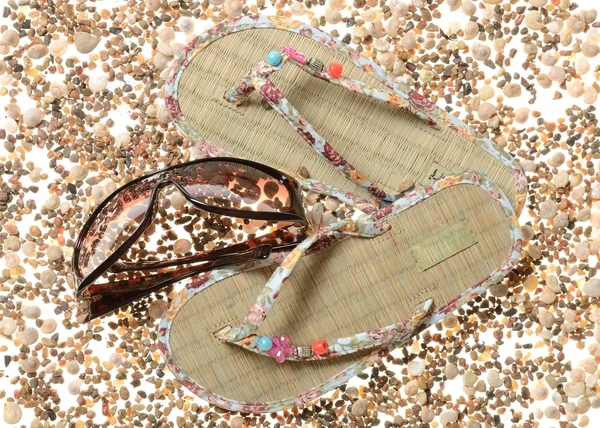 Braided barefoot persons sandals and women's sunglasses on a background of sea cockleshells and pebbles of stones — Stock Photo, Image