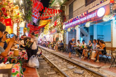 Turistler ve yerliler, Hanoi 'nin ünlü tren yolu boyunca kahve ve içkinin keyfini çıkarıyorlar. Renkli ışıklar ve şenlikli bir dekor ile parlıyorlar..
