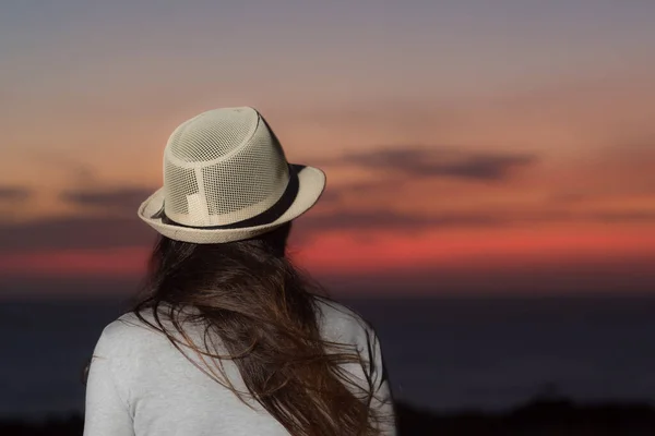 Close-up of woman with hat from her back looking at the sea at sunset.