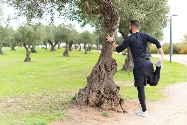 Joven corredor hombre con máscara estirándose en un parque apoyado contra un árbol —  Fotos de Stock