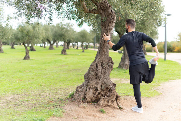 Молодой бегун в маске, растянувшись в парке, прислонился к дереву