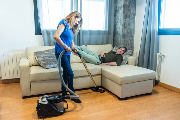 Kadın oturma odasını süpürürken adam kanepede yatıyor..