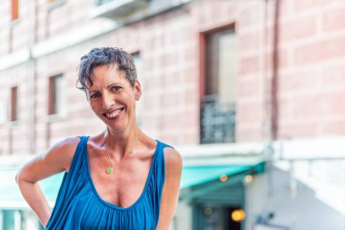 Caddede kameraya bakan neşeli, kısa saçlı olgun bir kadının portresi.