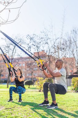Fitness çifti parkta trx fitness kayışlarıyla bacak egzersizi yapıyor.
