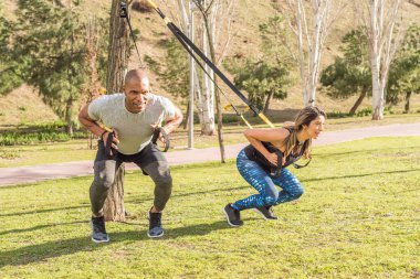 Athletic partners doing squatting back exercise with fitness straps in park