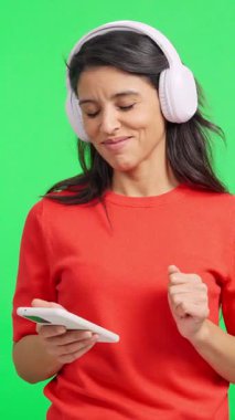 Kulaklıklı neşeli bir kadın müzik dinlemek ve yeşil ekranda dans etmek için akıllı telefon kullanıyor.