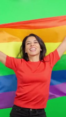 Gururlu gülümseyen kadın kendini gökkuşağı gurur bayrağına sarıp yeşil ekran arka planına yaslıyor.