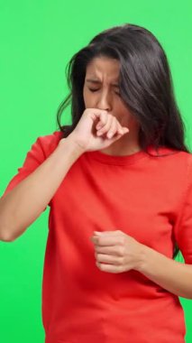 Sağlıksız genç bir kadın yeşil ekrana karşı öksürüyor ve ağrıyan boğazına dokunuyor.