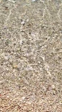 Güneşli bir günde çakıl taşlı bir sahil şeridinde süzülen yumuşak ve berrak su dalgalarının yukarıdan görünüşü