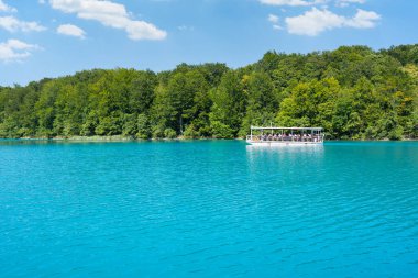 Turkuaz gölde mavi gökyüzünün altında yeşil ormanlarla çevrili turistler kayıkla geziyorlar.
