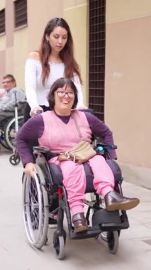 Genç bir kadın, dışarıda tekerlekli sandalyede fiziksel engelli mutlu bir orta yaşlı insanı itiyor.