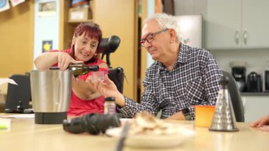 Tekerlekli sandalyede sakat gülümseyen bir kadın yemek pişirirken bir erkeğe zeytinyağı döküyor.