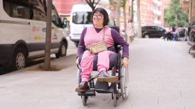 Şehrin kaldırımlarında dolaşırken tekerlekli sandalye kullanan, fiziksel engelli, mutlu yetişkin bir kadın.