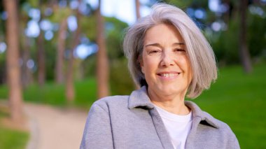 Şık kısa gri saçlı neşeli olgun kadın kameraya bakıyor ve parkta gülümsüyor.