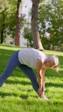 Yetişkin bir kadın, şehir parkındaki yeşil çimlerde antrenman yapmadan önce bacaklarını açıyor ve ısınıyor.