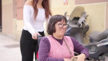 Bir arkadaşı tarafından tekerlekli sandalyeye itilirken gülümseyen fiziksel engelli genç bir kadın.