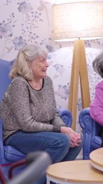 Bakıcı, bakım evindeki koltukta oturan yaşlı bir kadına rahatlık ve destek sağlıyor.