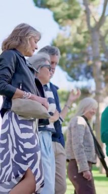 Neşeli, gri saçlı, güneşli bir parkta yürüyen ve konuşan bir grup yaşlı insan.