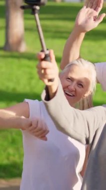Güneşli bir parkta birlikte video çekerken gülüp el sallayan bir grup yaşlı arkadaş.