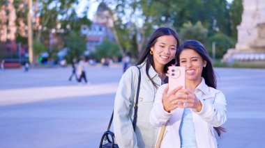 İki farklı kadın gülümsüyor, kaynaşıyor ve dışarıda akıllı bir telefonla selfie çekiyor.