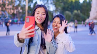 İki farklı kadın arkadaş gülümsüyor, el sallıyor ve cep telefonuyla video görüşmesi yapıyorlar.