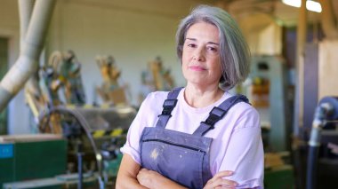 Olgun kadın operatör tulum giyiyor ve marangozluk atölyesinde kendinden emin görünüyor.