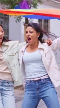 İki mutlu çok ırklı kadın bir geçit töreninde lgbtq artı bayrağıyla gülüyor ve yürüyorlar.