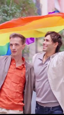 Genç, gülümseyen eşcinsel çift gururla yürüyor sokakta Gökkuşağı bayrağıyla