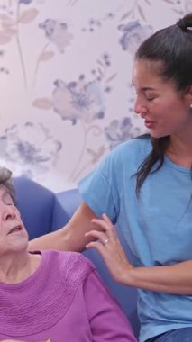 Genç bir kadın, yaşlı büyükannesini sevgiyle teselli ediyor. Bir bakım evinde bir sandalyede oturuyor.