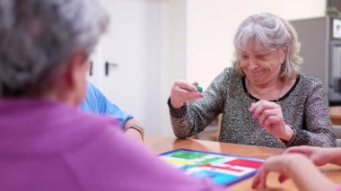Huzurevinde bakıcısıyla masa oyunu oynarken gülümseyen gri saçlı yaşlı bir kadın.