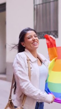 Elinde gökkuşağı bayrağıyla gururla yürüyen genç bir kadın ağır çekimde sokakta yürüyor.