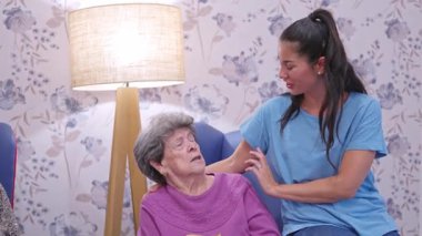 Genç bir kadın, yaşlı büyükannesini sevgiyle teselli ediyor. Bir bakım evinde bir sandalyede oturuyor.