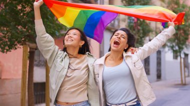 İki neşeli kadın, renkli bir gökkuşağı bayrağıyla yürüyorlar ve tezahürat yapıyorlar.