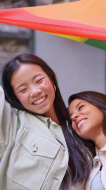 Birbirine aşık iki genç kadın gökkuşağı bayrağını tutarken açık havada sarılıp gülüyorlar.