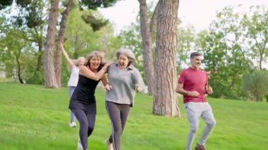 Bir grup mutlu kıdemli arkadaş, güneşli bir günde yeşil çimlerde neşeli bir şekilde koşuyorlar.
