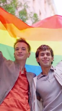 İki genç adam başlarının üzerinde gökkuşağı bayrağı tutarken kameraya bakıp gülümsüyor.