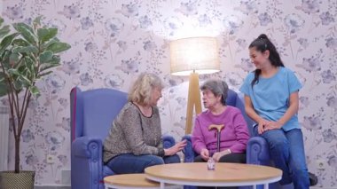 Yaşlı annesini huzurevinde ziyaret eden bir kız. Gülümseyen bir bakıcı ise onlara yardım ediyor.