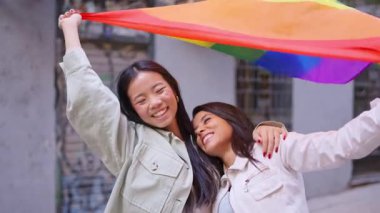 Birbirine aşık iki genç kadın gökkuşağı bayrağını tutarken açık havada sarılıp gülüyorlar.