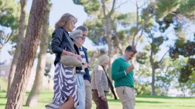Neşeli, gri saçlı, güneşli bir parkta yürüyen ve konuşan bir grup yaşlı insan.