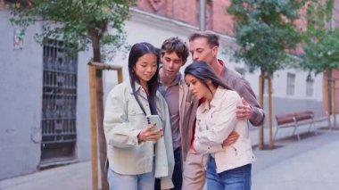 Çok ırklı bir arkadaş grubu açık havada gülümserken akıllı telefondan bir şey izliyor.