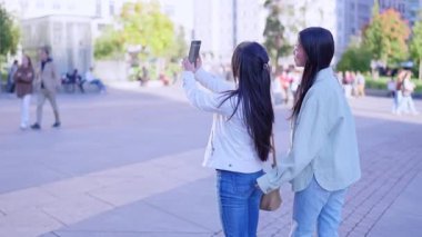 İki genç Asyalı kadın dışarıda akıllı bir telefonla selfie çekerken sarılıp gülümsüyor.