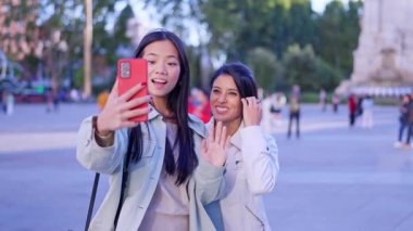 İki mutlu çok ırklı kadın dışarıda dolaşırken arkadaşlarıyla görüntülü konuşma yapmak için akıllı telefon kullanıyor.