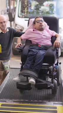Bakıcı, elektrikli tekerlekli sandalyedeki genç bir adama ulaşılabilir bir minibüs asansöründe yardım ediyor.
