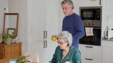 Yaşlı bir adam karısına portakal suyu getiriyor ve modern mutfaklarında kahvaltı yapıyor.